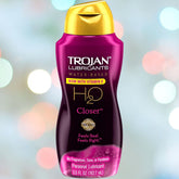 A bottle of Trojan H2O Closer - Water-Based Lubricant is set against a backdrop featuring colorful bokeh lights. This lubricant, enriched with vitamin E, is free from fragrances, dyes, or parabens and comes in a convenient 5.5oz size for enhanced intimacy.